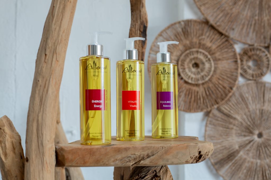 Trois flacons d'huiles de massage dorées aux étiquettes rouges et violettes disposés sur bois flotté pour rituel spa hiver